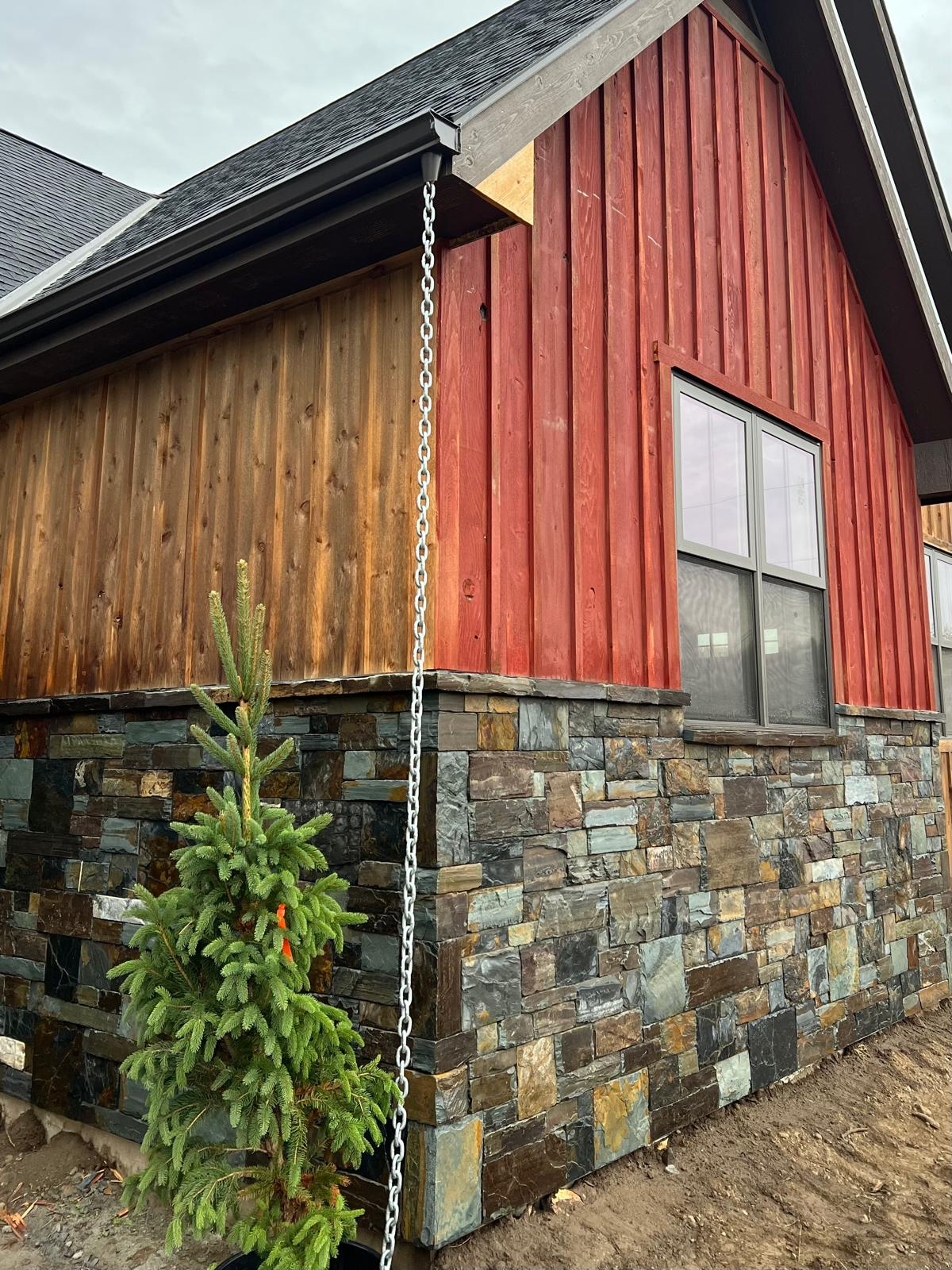 Alano Masonry stone veneer around a house corner with red and natural wood siding, a dark-framed window, a rain chain, and an evergreen